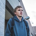 The image displays a sad teenager standing next to a school on an overcast day. He looks very tense and nervous. He is wearing a hoodie and a backpack.
